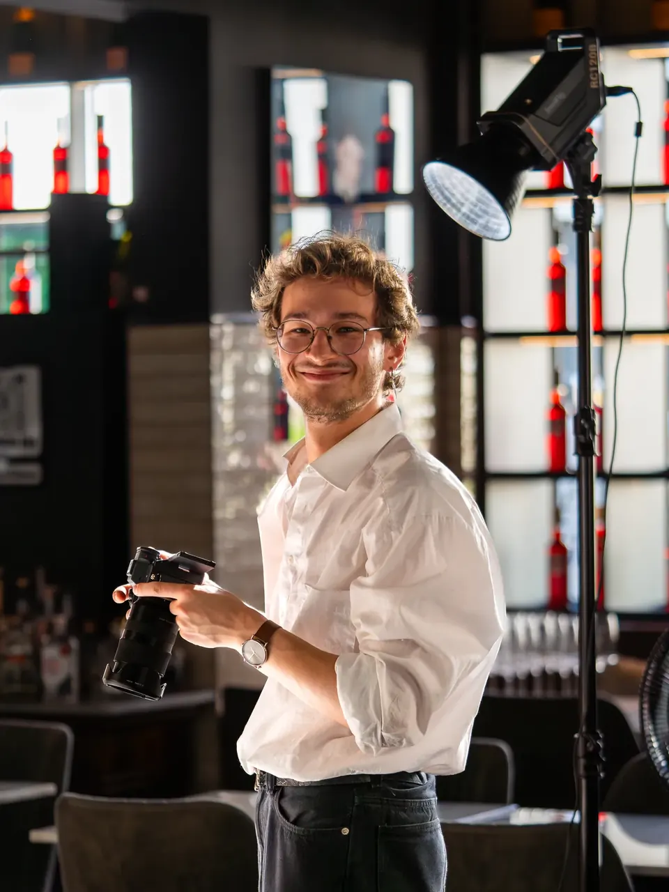 Videograf lächelt mit Kamera in der Hand neben der Studioleuchte in einer Bar