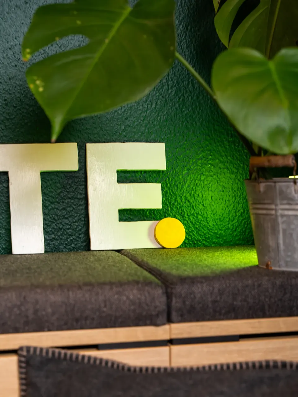 Buchstaben mit gelbem Punkt und Monstera vor der grünen Bürowand
